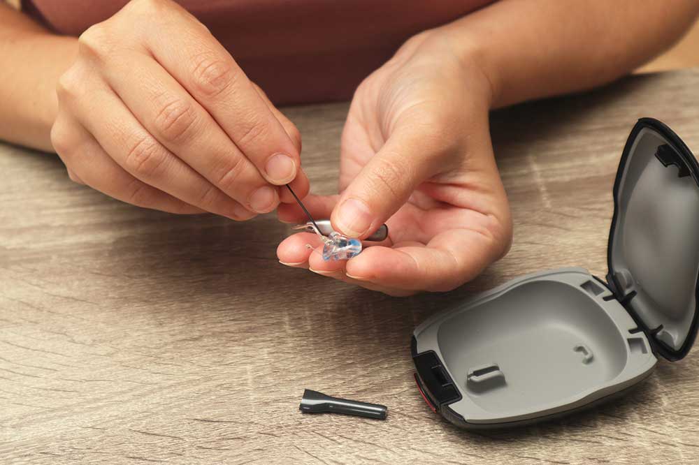 Hands using a wax loop on a hearing aid dome beside an open case, a cleaning step taught at Clear Choice Hearing Aid, Inc. in Southern California